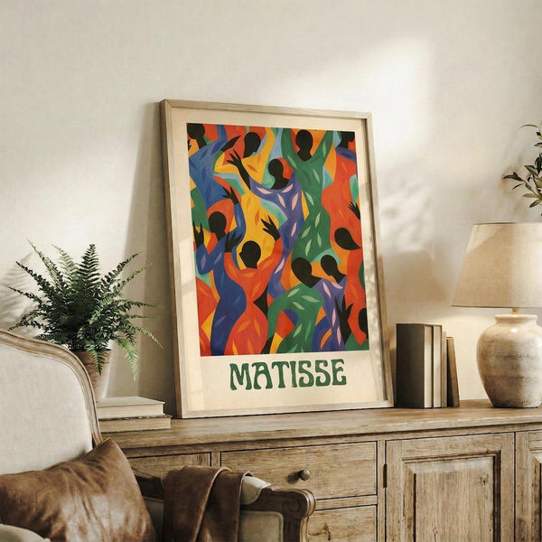 May include: Vibrant abstract art print featuring colorful dancing figures in blues, greens, oranges, and purples with black silhouetted heads, displayed in light wood frame on rustic wooden sideboard alongside fern plant, books, and ceramic vase in neutral-toned living room setting