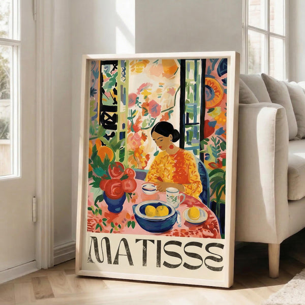May include: Colorful Matisse-style poster in light wood frame leaning against white wall next to beige couch. Artwork shows figure in yellow top at table with blue bowl, flowers, and tea setting surrounded by vibrant floral patterns in oranges, greens, and reds. Modern living room with hardwood floors and large windows.