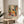 May include: Framed art print displaying colorful Matisse-style illustration of person in yellow shirt sitting at table with fruit bowl, teapot, and red flowers, surrounded by vibrant foliage and decorative patterns. Print rests on wooden round side table next to beige upholstered chair with palm plant visible in bright, minimalist room with white curtains and natural sunlight streaming across scene.