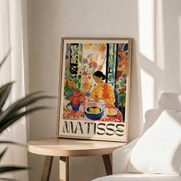 May include: Framed art print displaying colorful Matisse-style illustration of person in yellow shirt sitting at table with fruit bowl, teapot, and red flowers, surrounded by vibrant foliage and decorative patterns. Print rests on wooden round side table next to beige upholstered chair with palm plant visible in bright, minimalist room with white curtains and natural sunlight streaming across scene.