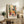 May include: Colorful Matisse art poster in light wood frame displayed on rustic wooden sideboard next to fern plant, books, and ceramic vase. Artwork features vibrant woman in yellow dress at table with red flowers and fruit bowls in bold fauve style with blues, greens, oranges and reds against cream matting.
