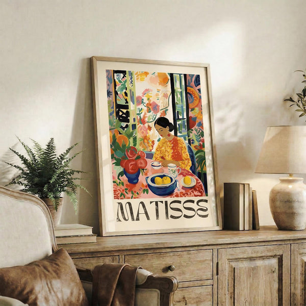 May include: Colorful Matisse art poster in light wood frame displayed on rustic wooden sideboard next to fern plant, books, and ceramic vase. Artwork features vibrant woman in yellow dress at table with red flowers and fruit bowls in bold fauve style with blues, greens, oranges and reds against cream matting.