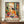 May include: Framed colorful Matisse-style poster featuring woman in yellow top sitting at table with fruit bowl, teacup, and red flowers by window. Vibrant fauve colors with bold brushstrokes. Frame sits on wooden sideboard next to white ceramic vase and stacked books in modern room with white paneled walls.