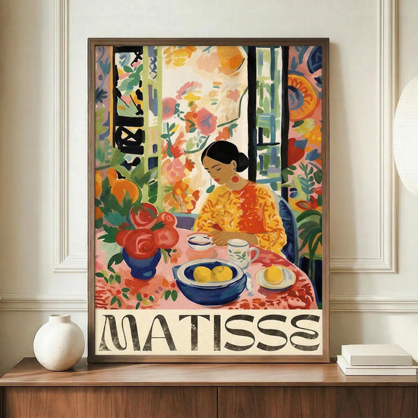 May include: Framed colorful Matisse-style poster featuring woman in yellow top sitting at table with fruit bowl, teacup, and red flowers by window. Vibrant fauve colors with bold brushstrokes. Frame sits on wooden sideboard next to white ceramic vase and stacked books in modern room with white paneled walls.