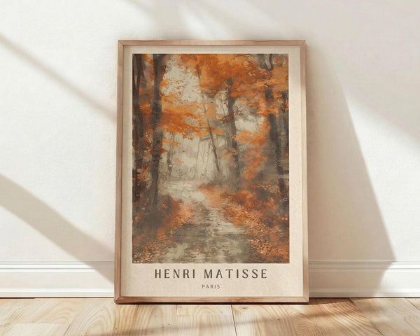 May include: Framed art print featuring an autumn forest scene with orange and brown foliage, dark tree trunks, and a misty pathway. The artwork shows painterly brushstrokes in warm earth tones with "HENRI MATISSE PARIS" text below the image. Natural wood frame sits against white wall on light wooden floor with soft sunlight shadows.