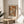 May include: Framed forest landscape poster with autumn orange and brown foliage, featuring "HENRI MATISSE PARIS" text below artwork. Print sits on light wood round side table next to beige upholstered chair and plant leaves, with natural sunlight creating shadows on cream wall background.