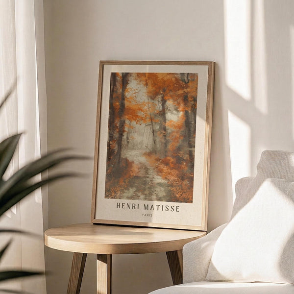 May include: Framed forest landscape poster with autumn orange and brown foliage, featuring "HENRI MATISSE PARIS" text below artwork. Print sits on light wood round side table next to beige upholstered chair and plant leaves, with natural sunlight creating shadows on cream wall background.