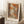 May include: Framed autumn forest scene print leaning against white wall corner, showing orange and brown foliage along a woodland path with dark tree trunks. Print has cream matting with "Henri Matisse Paris" text below artwork. Scene includes white coffee cup on wooden block and beige knitted blanket on textured white rug.