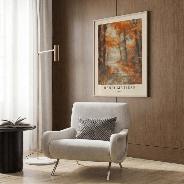 May include: Light beige armchair with geometric patterned pillow in modern room setting with warm wood paneling. Framed autumn forest artwork with orange and brown foliage hanging on wood wall, featuring brass floor lamp and dark side table with open book.