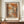 May include: Framed art print displaying an autumn forest scene with orange and brown foliage, dark tree trunks, and a misty path. The artwork features warm earth tones in an impressionistic style with "HENRI MATISSE PARIS" text below. Shown in thin dark frame on wooden sideboard next to white ceramic vase and stacked books against cream wainscoted wall.