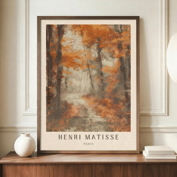 May include: Framed art print displaying an autumn forest scene with orange and brown foliage, dark tree trunks, and a misty path. The artwork features warm earth tones in an impressionistic style with "HENRI MATISSE PARIS" text below. Shown in thin dark frame on wooden sideboard next to white ceramic vase and stacked books against cream wainscoted wall.