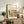 May include: Framed art poster with colorful abstract botanical design featuring trees, leaves, and organic shapes in vibrant yellows, greens, blues, and oranges. Text reads "Discover the beauty of Matisse's art" above the artwork. Natural wood frame displayed on rustic wooden sideboard alongside fern plant, ceramic vases, books, and beige upholstered chair in modern living room setting with neutral walls.