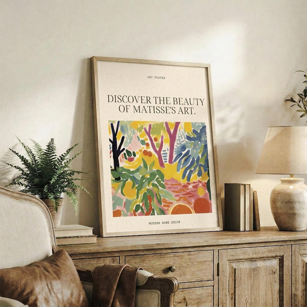 May include: Framed art poster with colorful abstract botanical design featuring trees, leaves, and organic shapes in vibrant yellows, greens, blues, and oranges. Text reads "Discover the beauty of Matisse's art" above the artwork. Natural wood frame displayed on rustic wooden sideboard alongside fern plant, ceramic vases, books, and beige upholstered chair in modern living room setting with neutral walls.