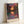 May include: Framed art print leaning against white wall showing vibrant landscape with trees silhouetted against colorful sunset sky reflected in water below. Artwork features bold purples, oranges, greens and pinks in impressionist style. Light wooden frame with "HENRI MATISSE PARIS" text at bottom. Natural lighting creates soft shadows on wall.