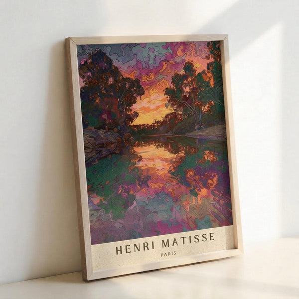 May include: Framed art print leaning against white wall showing vibrant landscape with trees silhouetted against colorful sunset sky reflected in water below. Artwork features bold purples, oranges, greens and pinks in impressionist style. Light wooden frame with "HENRI MATISSE PARIS" text at bottom. Natural lighting creates soft shadows on wall.