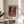 May include: Framed art print featuring a colorful landscape with trees reflected in water, displayed on a light wood round side table next to a beige upholstered chair. The artwork shows vibrant purples, oranges, and greens in an impressionist style with "HENRI MATISSE PARIS" text below. Natural sunlight creates shadows on the cream-colored wall behind.