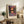 May include: Framed art print displaying a vibrant landscape with colorful trees reflected in water, featuring rich purples, oranges, and greens in an impressionistic style. The print sits on a rustic wooden sideboard alongside books, a ceramic vase, and a fern plant, with a cream upholstered chair visible in a warmly lit living room setting.