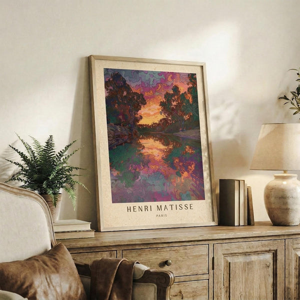 May include: Framed art print displaying a vibrant landscape with colorful trees reflected in water, featuring rich purples, oranges, and greens in an impressionistic style. The print sits on a rustic wooden sideboard alongside books, a ceramic vase, and a fern plant, with a cream upholstered chair visible in a warmly lit living room setting.