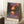 May include: Framed art print leaning against white wall showing vibrant landscape with trees, water reflection, and colorful sky in purple, orange, and green tones. Print features artist name at bottom. Scene includes coffee cup on wooden surface and textured white rug.