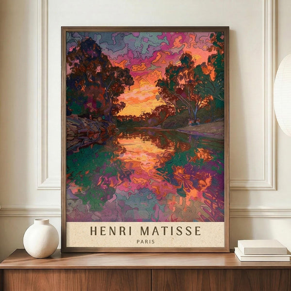 May include: Framed art print featuring a vibrant landscape with trees surrounding a reflective river at sunset, rendered in bold colors including pink, orange, purple, and green with distinctive outlined forms. The poster displays on a wooden sideboard next to a white ceramic vase and stacked books in an elegant room with cream walls and white molding.