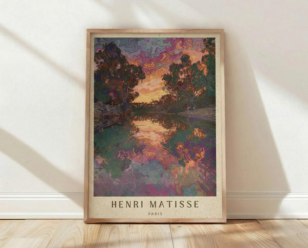 May include: Framed art print showing a vibrant landscape with swirling purple and pink clouds above dark trees flanking a reflective stream. The colorful scene features rich purples, oranges, and greens in an expressive painterly style. The print sits in a light wood frame against white walls on blonde hardwood flooring, with soft natural light creating diagonal shadows across the wall.