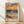 May include: Framed poster print featuring an impressionist seascape painting with vibrant orange sunset sky, turbulent blue-green ocean waves, and sandy beach with tall grass in foreground. The artwork shows expressive brushstrokes in warm oranges, cool blues, and earth tones. Text reading "MATISSE" appears below the image. Light wood frame sits against white textured wall on natural wood flooring with soft natural lighting casting shadows.