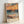 May include: Framed art print featuring a vibrant seascape painting in an impressionist style, displayed in a light wooden frame leaning against a white wall. The artwork shows rolling ocean waves in blues and teals, with a dramatic sunset sky in warm oranges and corals above. Beach grass appears in the foreground, and "MATISSE" text is printed at the bottom of the piece.