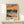 May include: Framed poster print displayed on light wood console against cream wall. Artwork shows vibrant seascape with dramatic orange and coral sunset sky, teal ocean waves, and sandy beach with grass in foreground. Bold brushstroke style with "MATISSE" text at bottom. Natural wood frame, small wrapped gift box beside it.