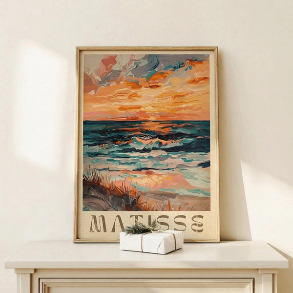 May include: Framed poster print displayed on light wood console against cream wall. Artwork shows vibrant seascape with dramatic orange and coral sunset sky, teal ocean waves, and sandy beach with grass in foreground. Bold brushstroke style with "MATISSE" text at bottom. Natural wood frame, small wrapped gift box beside it.