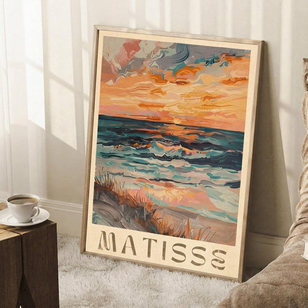 May include: Framed coastal landscape poster with vibrant orange sunset sky and teal ocean waves, featuring thick painterly brushstrokes in an impressionist style. The artwork shows a beach scene with marsh grass in the foreground and "MATISSE" text at the bottom. Displayed in a light wood frame leaning against white walls in a cozy room setting with a coffee cup on a wooden side table and soft beige blanket nearby.