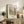 May include: Framed beige and black sketch artwork showing a peaceful park scene with trees, water, and pathways displayed on a rustic wooden sideboard alongside decorative fern plant, stacked books, ceramic vase, and table lamp in a cozy living room setting with neutral tones and natural lighting.
