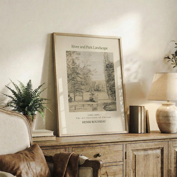 May include: Framed beige and black sketch artwork showing a peaceful park scene with trees, water, and pathways displayed on a rustic wooden sideboard alongside decorative fern plant, stacked books, ceramic vase, and table lamp in a cozy living room setting with neutral tones and natural lighting.