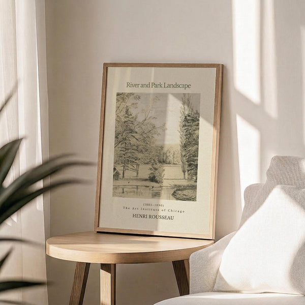 May include: A framed vintage landscape print displayed on a light wood round side table next to a beige upholstered chair. The artwork shows a black and white riverside park scene with trees, featuring text at the top and bottom. Natural sunlight creates soft shadows across the neutral-toned room setting with a plant visible in the corner.