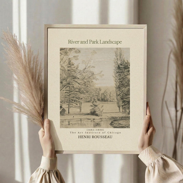 May include: Person in cream sweater holding framed vintage landscape print showing pastoral river scene with trees, small buildings, and water. Black and white sketch-style artwork in beige matted frame. Dried wheat stalks visible on left side. Soft natural lighting against neutral curtained background.