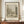 May include: Framed vintage-style poster displaying a sepia-toned pencil sketch of a peaceful riverside scene with trees, small buildings, and water reflections. The artwork features "River and Park Landscape" title, dates (1885-1890), "The Art Institute of Chicago" and "HENRI ROUSSEAU" text below the illustration. Displayed on a warm wood credenza with white ceramic vase and stacked books against cream walls with white trim molding.