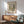 May include: Framed vintage Henri Rousseau landscape print hanging on white wall above wood sideboard with table lamp. Green mid-century armchair with wooden arms sits nearby. Artwork shows sepia-toned winter river scene with bare trees, bridge, and snow. Warm sunlight casts shadows across the minimalist room setting with round coffee table.