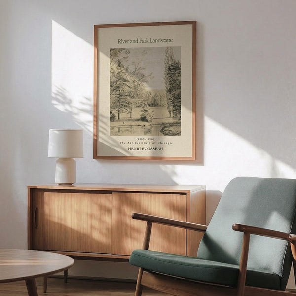May include: Framed vintage Henri Rousseau landscape print hanging on white wall above wood sideboard with table lamp. Green mid-century armchair with wooden arms sits nearby. Artwork shows sepia-toned winter river scene with bare trees, bridge, and snow. Warm sunlight casts shadows across the minimalist room setting with round coffee table.