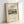 May include: Framed art print featuring Henri Rousseau's pastoral landscape painting with houses, trees, and winding path in muted greens and earth tones. White matted poster displays "SAWMILL OUTSKIRTS OF PARIS" text and museum details. Light wood frame leans against white wall corner, casting soft shadows. Clean minimalist styling showcases vintage exhibition poster format.