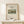 May include: Framed art print featuring Henri Rousseau's Sawmill painting with rustic buildings and green landscape, displayed on light wood shelf beside small wrapped gift with twine and pine sprig. Print has cream background with black text and natural wood frame against white wall.