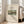 May include: Framed art print leaning against white wall next to beige upholstered chair. Print features pastoral landscape painting with green rolling hills, white buildings with red roofs, winding dirt path, and bare trees under pale sky. White matted frame with cream background and black text reading "SAWMILL OUTSKIRTS OF PARIS" and "Henri Rousseau" with museum details. Natural sunlight streams across hardwood floor in bright, minimalist living room setting.