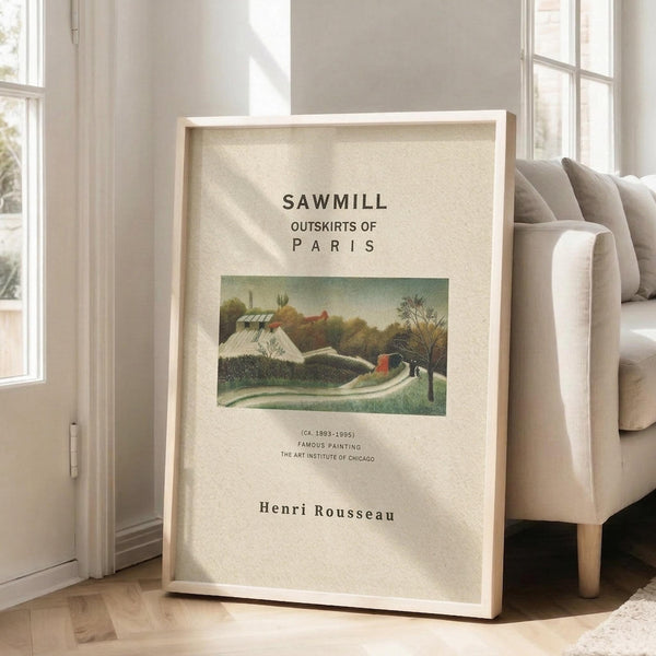 May include: Framed art print leaning against white wall next to beige upholstered chair. Print features pastoral landscape painting with green rolling hills, white buildings with red roofs, winding dirt path, and bare trees under pale sky. White matted frame with cream background and black text reading "SAWMILL OUTSKIRTS OF PARIS" and "Henri Rousseau" with museum details. Natural sunlight streams across hardwood floor in bright, minimalist living room setting.