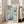 May include: A framed art print with blue and teal floral patterns and dots, featuring "Yayoi Kusama" text and "1972" at the bottom. The light wood frame leans against a white wall next to a beige sofa in a bright room with large windows and natural lighting.