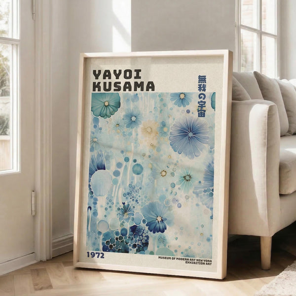 May include: A framed art print with blue and teal floral patterns and dots, featuring "Yayoi Kusama" text and "1972" at the bottom. The light wood frame leans against a white wall next to a beige sofa in a bright room with large windows and natural lighting.