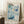 May include: Framed poster with blue and teal floral and bubble pattern artwork leaning against white wall corner. Print features "YAYOI KUSAMA" text at top, Japanese characters, and "1972 MUSEUM OF MODERN ART NEW YORK" at bottom. Light wood frame, displayed on white textured rug next to wooden side table with white coffee cup and saucer. Natural lighting creates soft shadows on wall.