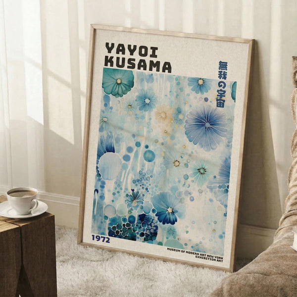 May include: Framed poster with blue and teal floral and bubble pattern artwork leaning against white wall corner. Print features "YAYOI KUSAMA" text at top, Japanese characters, and "1972 MUSEUM OF MODERN ART NEW YORK" at bottom. Light wood frame, displayed on white textured rug next to wooden side table with white coffee cup and saucer. Natural lighting creates soft shadows on wall.