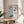 May include: Framed exhibition poster with blue and white abstract floral design featuring scattered circular patterns and flower motifs, displayed on light wood round side table next to beige upholstered chair, with plant leaves visible in corner and natural window lighting casting shadows on cream wall