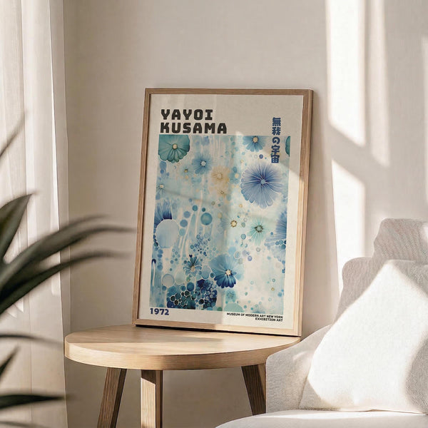 May include: Framed exhibition poster with blue and white abstract floral design featuring scattered circular patterns and flower motifs, displayed on light wood round side table next to beige upholstered chair, with plant leaves visible in corner and natural window lighting casting shadows on cream wall
