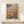 May include: Framed art print featuring colorful polka-dotted spherical sculptures on stems in orange, pink, and yellow tones against a muted background. The poster displays "YAYOI KUSAMA MUSEUM" text with Japanese characters. Set in a minimalist room scene with light wood frame, positioned on a white wooden surface next to a small wrapped gift box with natural twine and greenery accent.
