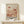 May include: Framed art print in natural wood frame featuring abstract botanical illustration with coral and terracotta mushrooms, dotted patterns, and organic shapes in warm earth tones. Text reads "YAYOI KUSAMA" and "KYOTO" in black letters. Print leans against white wall on light wood surface next to small wrapped gift box with twine and greenery sprig.