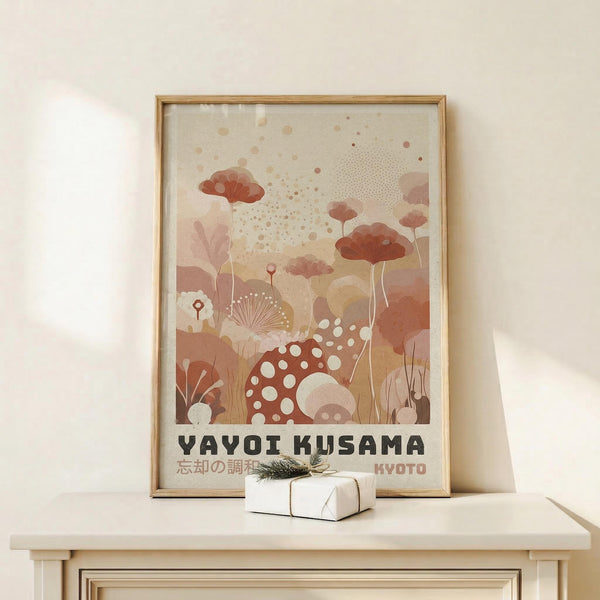 May include: Framed art print in natural wood frame featuring abstract botanical illustration with coral and terracotta mushrooms, dotted patterns, and organic shapes in warm earth tones. Text reads "YAYOI KUSAMA" and "KYOTO" in black letters. Print leans against white wall on light wood surface next to small wrapped gift box with twine and greenery sprig.